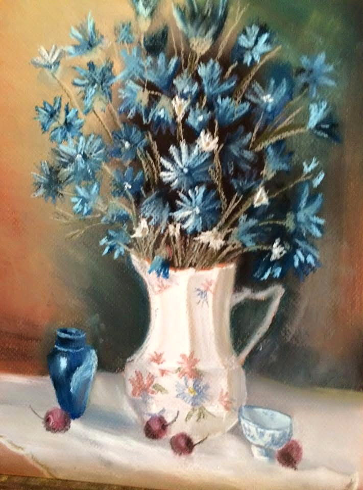 Blue wildflowers arranged in a white floral pitcher beside a small blue vase, teacup, and scattered fruit