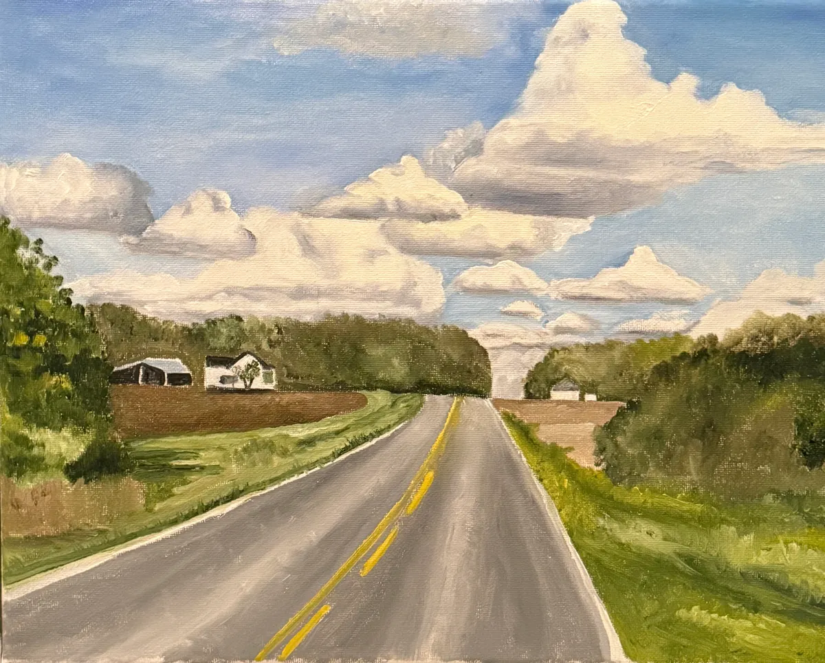 Straight rural highway beneath towering white clouds, fields, and farm buildings