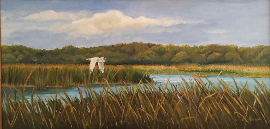 A white egret flying over blue marsh water and tall reeds beneath a cloud-filled sky at Meaher Park